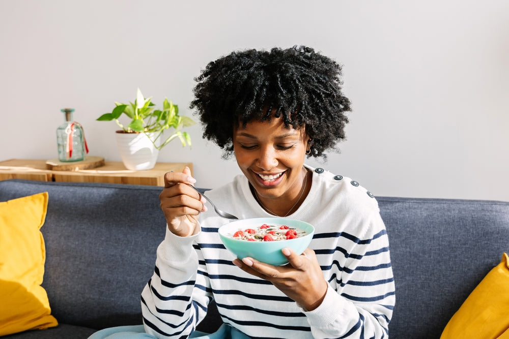 Happy young woman eating healthy breakfast bowl of yogurt and fruit sitting on sofa in the living room of her modern apartment. Healthy lifestyle and eating 